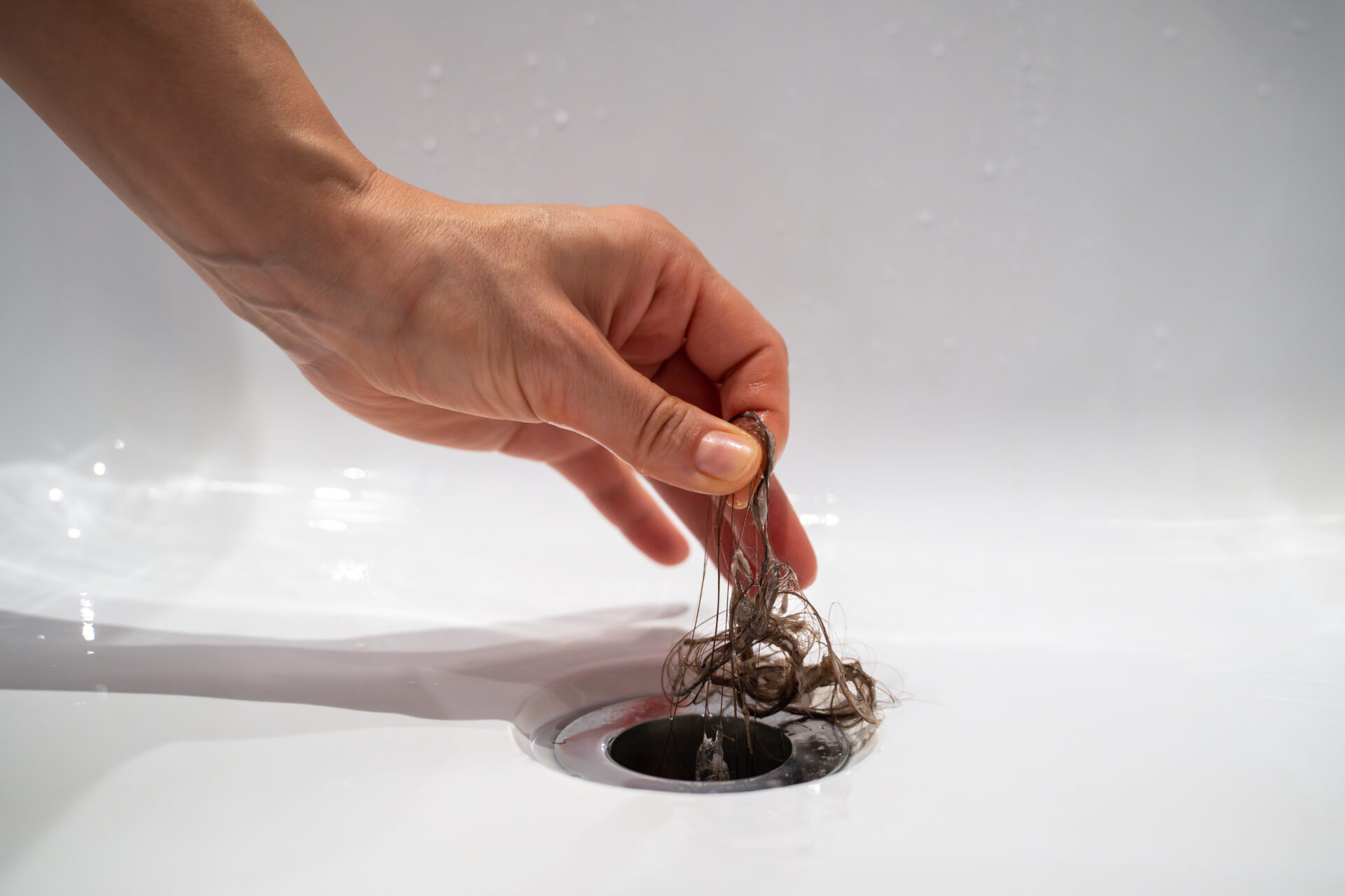 Water-drain-hole-in-bath-is-clogged-with hair-clump-in-bathroom-at-home. Female-cleaning-sewer-trap-taking-hairs-hand-closeup. Bath-plug-hole-blocked-with-dirt. Problem-with-shower-sewerage.